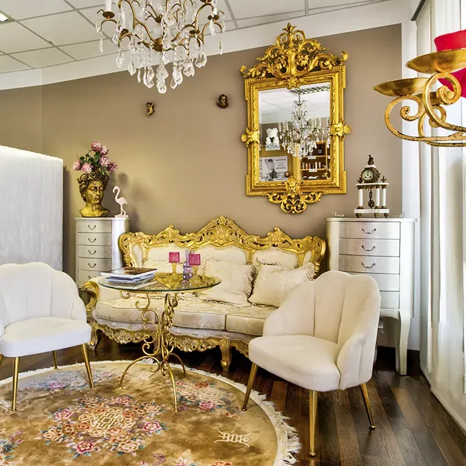 Der stilvoll eingerichteter Empfangs- oder Wartebereich von Julia Salchegger Kosmetikstudio mit luxuriösem, barockem Flair. Im Zentrum steht ein goldfarbenes Sofa mit beigefarbenen Polstern vor einem runden Glastisch, flankiert von zwei cremefarbenen Sesseln auf einem runden, gemusterten Teppich. Über dem Sofa hängt ein prunkvoller goldener Spiegel, ergänzt durch einen Kronleuchter. Im Raum befinden sich zudem elegante Kommoden, Dekorationselemente und ein Aufsteller der Marke „Dalton Marine Cosmetics“.