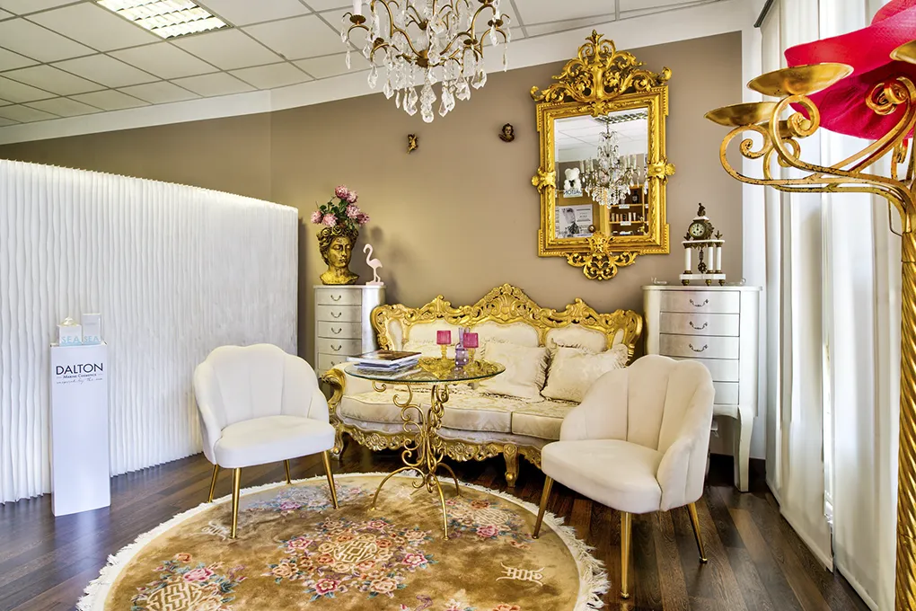Der stilvoll eingerichteter Empfangs- oder Wartebereich von Julia Salchegger Kosmetikstudio mit luxuriösem, barockem Flair. Im Zentrum steht ein goldfarbenes Sofa mit beigefarbenen Polstern vor einem runden Glastisch, flankiert von zwei cremefarbenen Sesseln auf einem runden, gemusterten Teppich. Über dem Sofa hängt ein prunkvoller goldener Spiegel, ergänzt durch einen Kronleuchter. Im Raum befinden sich zudem elegante Kommoden, Dekorationselemente und ein Aufsteller der Marke „Dalton Marine Cosmetics“.
